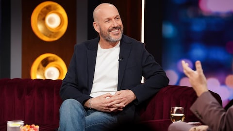 Martin Matte est assis sur un canapé et il sourit.