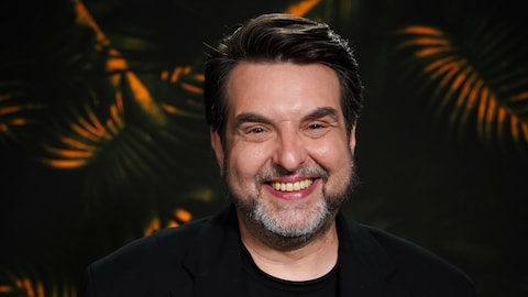 L'animateur et chroniqueur sourit à la caméra devant un décor tropical.
