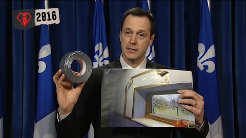 L'ancien ministre de l'éducation, Jean-François Roberge, tient un rouleau de ruban adhésif et une photo d'une salle de classe en décrépitude.