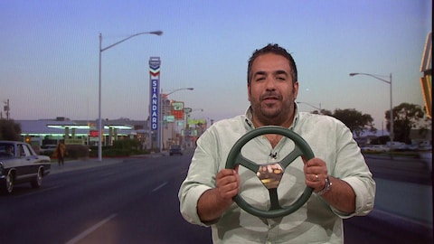 L'humoriste Neev tient un volant dans un faux décor de boulevard de banlieue.