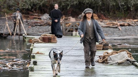 Un enfant marche sur un quai de bois et tient un chien en laisse. Un religieux suit loin derrière.
