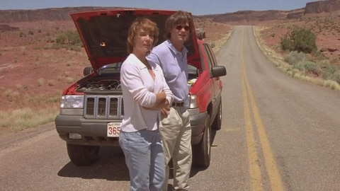 Un homme et une femme, devant une voiture le capot ouvert, sur une route dans le désert.