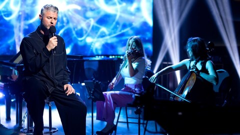 Pierre Lapointe chante en tenant un micro. Il est accompagné de deux musiciennes.