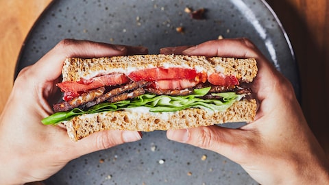 Deux mains tiennent un sandwich BLT au tempeh au-dessus d'une assiette avec un verre d’eau.