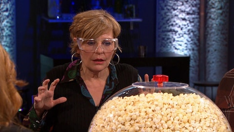 Elle porte des lunettes de sécurité et devant elle, se trouve un gros ballon en verre rempli de maïs soufflé. 