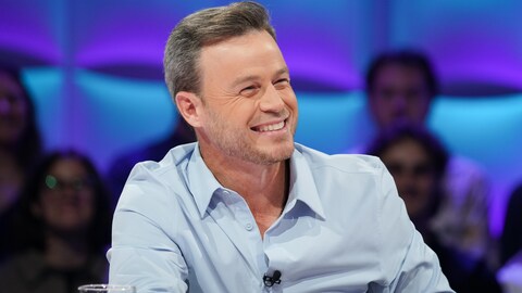 Il est assis en studio et rit.
