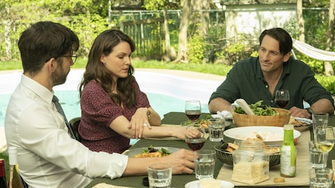 Les trois personnes sont assises à une table à l'extérieur avec de la nourriture et du vin. Les deux hommes se regardent.