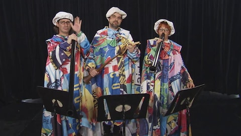 Zone 3 Jean-René Dufort, Mc Gilles et Chantal Lamarre sont sur scène déguisés avec des costumes artistiques extravagants et de faux instruments de musique.