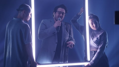 Bryan Audet chante avec un micro derrière un cadre fait de néons.