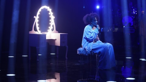 Naadei chante dans un micro assise sur un tabouret devant un miroir illuminé.