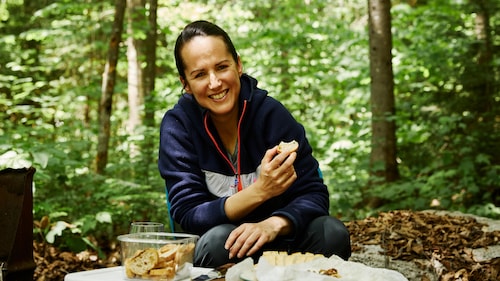 Geneviève O'Gleman en camping qui déguste des croutons de pain avec un fromage cuisiné sur un feu de camp.