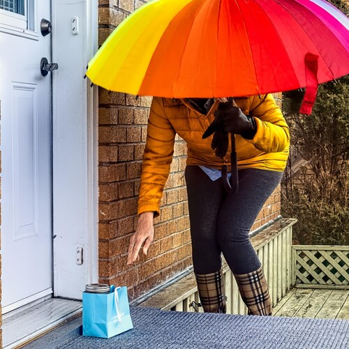 Devant le perron d'une maison,  à l'abri sous un parapluie, une personne est penchée pour prendre  un sac cadeau contenant de la nourriture.
