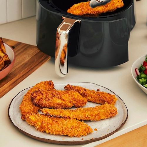Des frites, des doigts de poulet panés, une salade verte et le tiroir d’une friteuse à air sur le comptoir d’une cuisine. Des pinces tiennent un doigt de poulet.