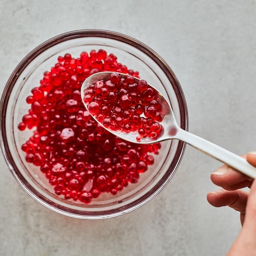 Une cuillère contenant des perles de jus de fruits au-dessus d'un bol en verre contenant des perles de jus de fruits.