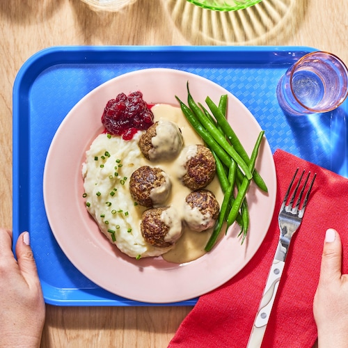 Un plateau de cafétéria avec une assiette contenant de la purée de pommes de terre, des boulettes suédoises et des haricots verts.