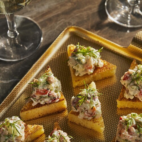 Plusieurs triangles de pain grillé recouverts d’une préparation au homard et d’une feuille d’aneth fraîche dans un plateau doré avec deux coupes de vin blanc.