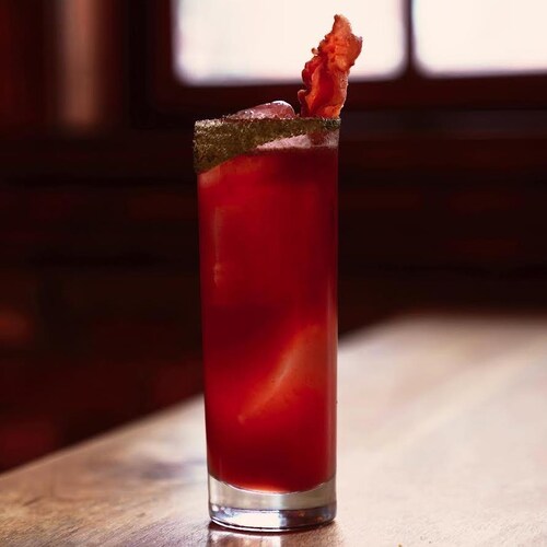 Un cocktail rouge dans un verre allongé avec une tranche de bacon en décoration.