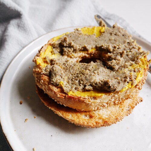 Une moitié de bagel grillé tartiné de de moutarde et de cretons végétaliens dans une assiette.