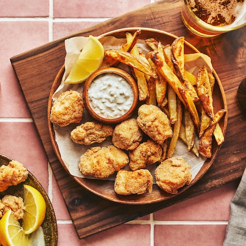 Des bouchées de fish and chips dans une assiette en bois sur une planche de bois, accompagnés de frites, d’une sauce tartare et de verres de bière. 