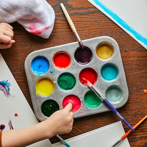 Plusieurs couleurs de gouache maison sont dans les cavités d’un moule à muffins. Un petit garçon trempe ses doigts dans la gouache, alors qu’une petite fille dessine un papillon avec un pinceau. 