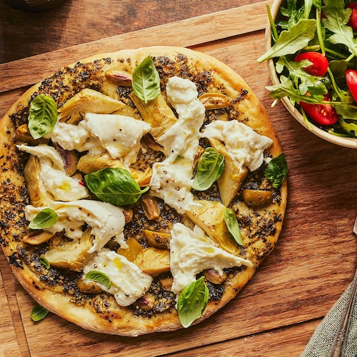 Sur une planche en bois, il y a une pizza aux artichauts et à la burrata et un bol de salade.