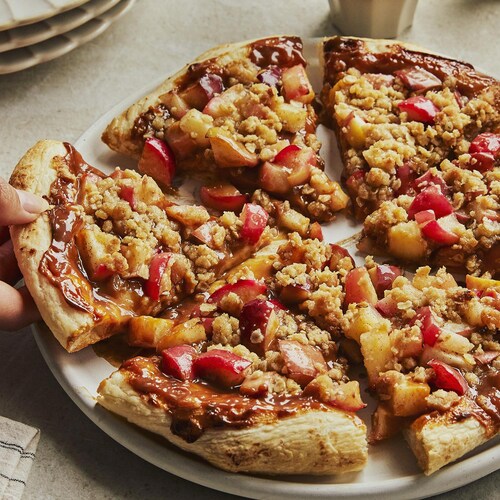 Dans une grande assiette, une main prend une pointe de la pizza dessert aux pommes. Autour, il y a une pile d’assiette et une petite tasse de café.