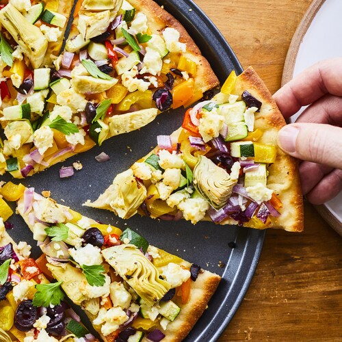 Une pizza garnie de coeurs d'artichauts, d'olives, de poivrons et d'oignons rouges sur une plaque cuisson avec une main qui prend une pointe.