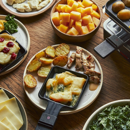 Au centre d’une table, il y a une assiette avec des patates grillées, des petits morceaux de porc cuit et une coupelle à raclette remplie de kale et de rutabaga gratinés. Autour, il y a un poêle à raclette, un bol de kale, une assiette de fromage, une assiette avec des patates gratinées et un bol de rutabaga en cube.