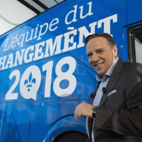 François Legault sourit pour les caméras alors qu'il se trouve devant l'autobus de la CAQ, sur laquelle est inscrit « L'équipe du changement 2018 ». 