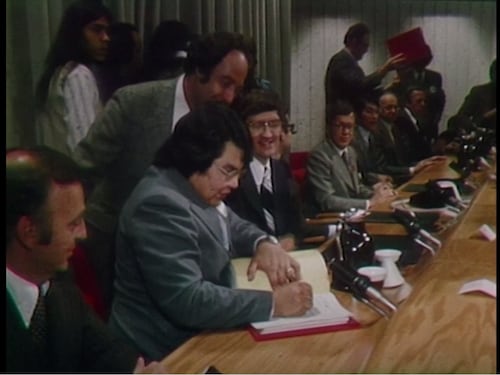 Homme qui signe un document sous le regard des autres hommes assis près de lui.