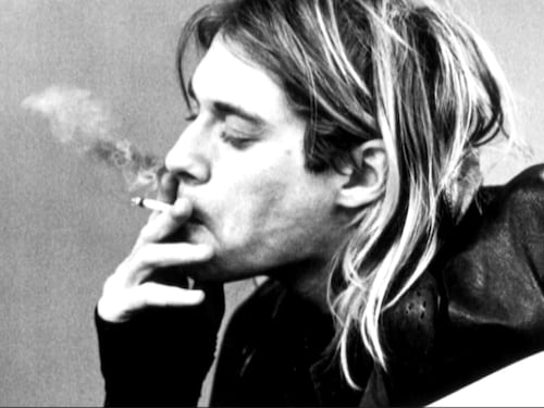 Kurt Cobain fume une cigarette appuyé sur sa guitare. 