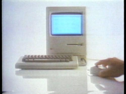 Écran et clavier du nouvel ordineur de Macintosh en 1984. Une main est posée sur la souris.