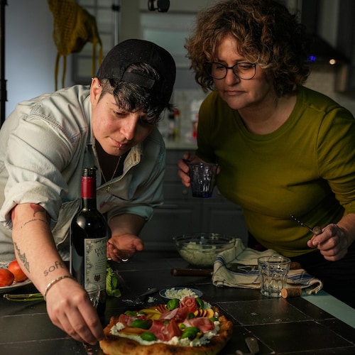 Dans une cuisine, Zoe Rousseau émiette avec soin une croûte de pizza sous le regard attentif de Tracy Cox. Sur le comptoir, il y a une bouteille de vin et une pizza recouverte de charcuteries, de basilic frais et de fruits.
