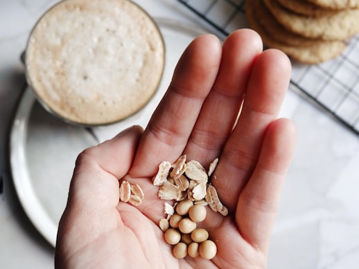 Des grains de soya et d'avoine dans le creux d'une main, et un verre de boisson moussée pour le café et des biscuits vus de haut, en arrière-plan.