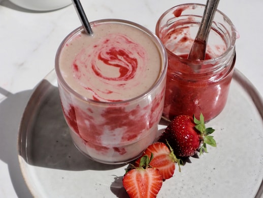 Une verre de smoothie à la fraise, un pot de coulis de fraises et trois fraises dans une assiette.