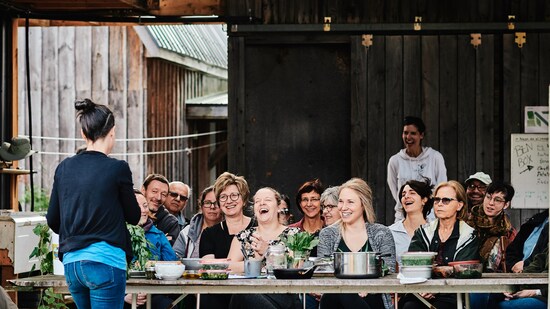 Une personne donne un atelier culinaire devant un groupe de gens assis derrière de longues tables en bois. 