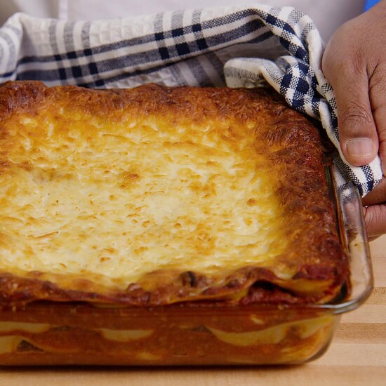 Une personne tient un plat de verre carré contenant une lasagne sortie du four. 