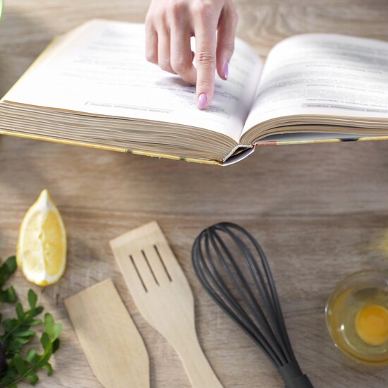 Les mains d'une femme qui lit un livre de recette posé sur un comptoir de cuisine, avec des ingrédients et des instruments.