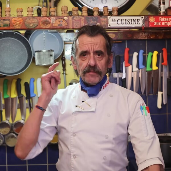 Le chef regarde dans l'objectif de la caméra derrière son comptoir de cuisine. Derrière lui, on voit plusieurs casseroles accrochées à des crochets au plafond et des couteaux accrochés à une barre magnétique pour couteaux.