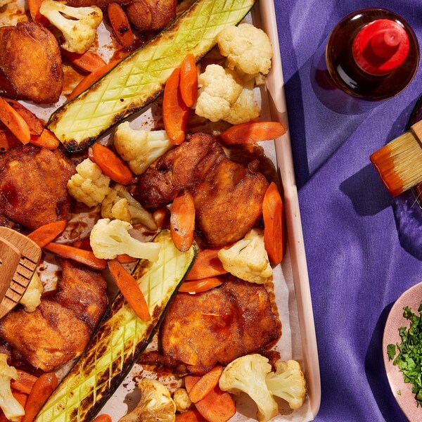 Des hauts de cuisses de poulet laqués de sauce BBQ, des tranches de carotte, des fleurons de chou-fleur et des courgettes sur une plaque à cuisson.
