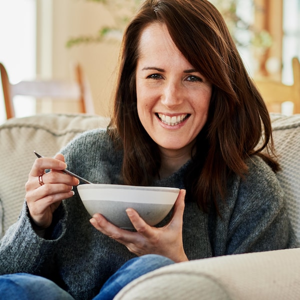 Geneviève O'Gleman tient un bol de soupe dans ses mains. Elle assise dans un fauteuil en tissu moelleux.
