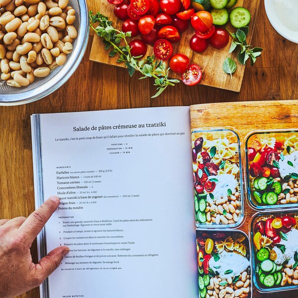 Un livre de recettes ouvert sur un plan de travail entouré de haricots blancs, de tomates cerise et de pâtes alimentaires.