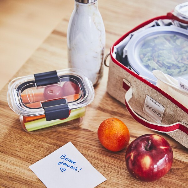 Un contenant avec des crudités, une clémentine, une pomme et une boîte à lunch ouverte sont sur une table, accompagnés d’un mot sur lequel est écrit bonne journée. 