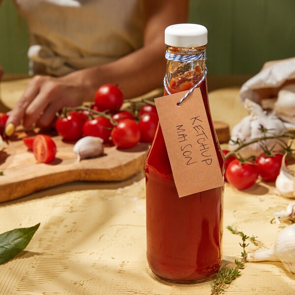 Une bouteille remplie de ketchup maison et une femme qui tranche des tomates en arrière-plan.