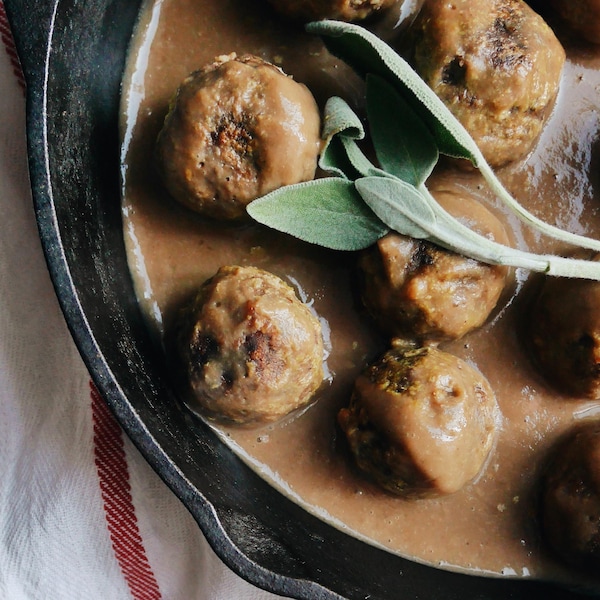 Vue en plongée sur une demi-poêle en fonte contenant des boulettes baignant dans une sauce et garnies de feuilles de sauge.