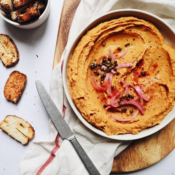 Un bol de tartinade garnie d'oignons et de câpres, un couteau, trois croûtons et un pot d'ingrédients posés sur une table.