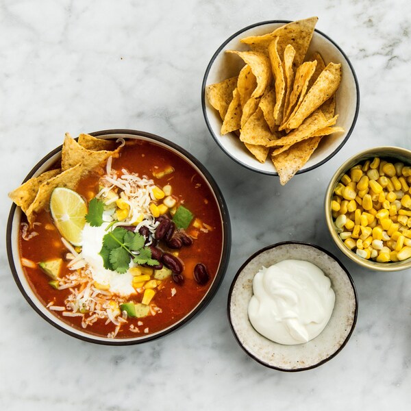 Un bol de soupe garni, plusieurs bols contenants des grains de maïs, des haricots rouge, du fromage râpé, des croustilles de maïs, de la crème sure, de la coriandre.