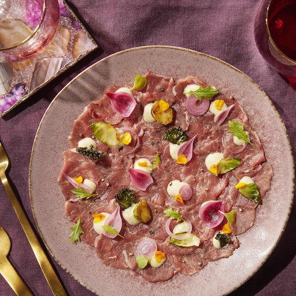 Dans une assiette, des tranches minces d'agneau sont disposées en rond. Le carpaccio est accompagné de feuilles de chou de Bruxelles, de chips de kale ainsi que des gouttes de sauce ranch.
