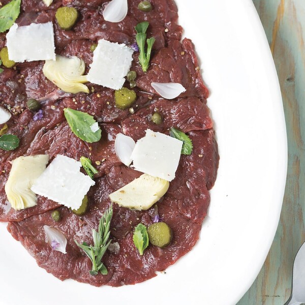 Une assiette ovale contient des tranches fines de viande d'orignal crues, étalées sur toute l'assiette et garnies de fines herbes.