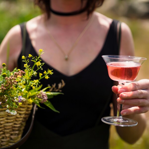 Une femme tenant un panier de fleurs sauvages dans une main et un cocktail dans l'autre. 